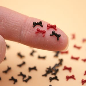 May include: A close-up of a finger holding several small black and red bows. The bows are made of plastic and are about the size of a dime. There are many more bows scattered on a light brown surface.