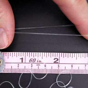 May include: A close-up of a clear elastic string being stretched between two fingers. A pink ruler with black markings is below the string, showing measurements in inches and centimeters. The ruler reads "1 60 in 2 3 cm".