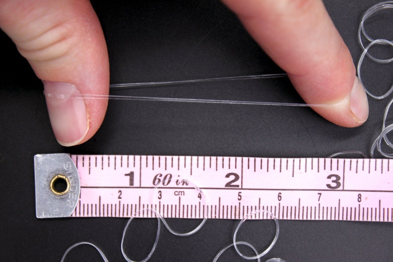 May include: A close-up of a clear elastic string being stretched between two fingers. A pink ruler with black markings is below the string, showing measurements in inches and centimeters. The ruler reads "1 60 in 2 3 cm".