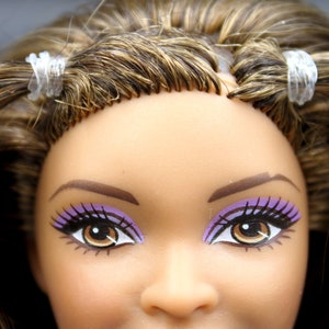 May include: Close-up of a doll's face with brown hair, purple eyeshadow, and brown eyes. The doll's hair is pulled back with clear hair ties.