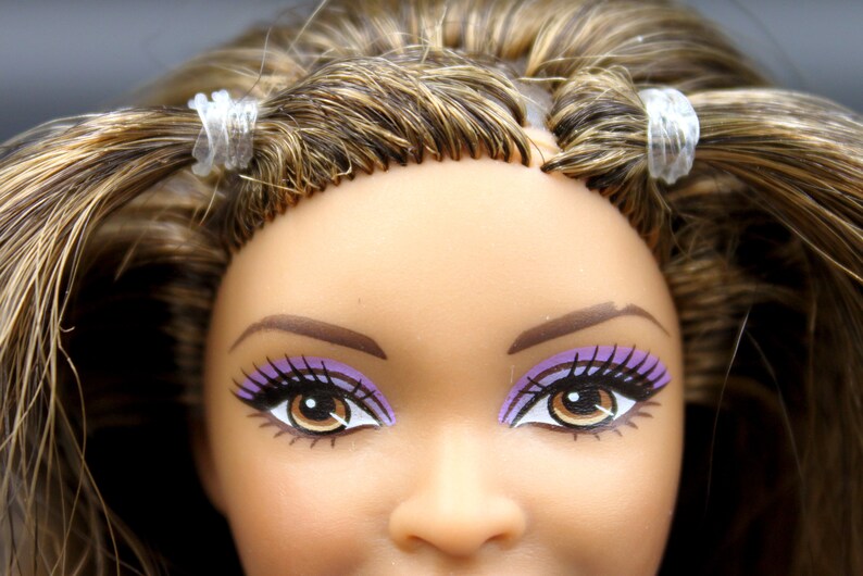 May include: Close-up of a doll's face with brown hair, purple eyeshadow, and brown eyes. The doll's hair is pulled back with clear hair ties.