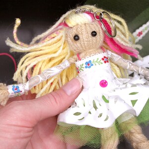 May include: A handmade yarn doll with long blonde hair, pink and yellow yarn accents, and a white lace dress with floral embroidery. The doll is wearing a green tulle skirt and has a key ring attached to its head.