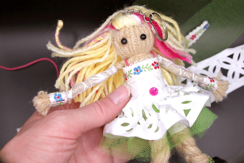 May include: A handmade yarn doll with long blonde hair, pink and yellow yarn accents, and a white lace dress with floral embroidery. The doll is wearing a green tulle skirt and has a key ring attached to its head.