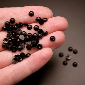 May include: A pile of black plastic buttons in a hand. The buttons are round and have a flat back.