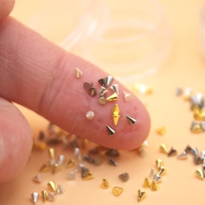 May include: Close-up of various metallic nail art studs in gold, silver, and bronze tones. The studs are cone-shaped and diamond-shaped, scattered on a finger and a light orange surface. Small containers are in the background.