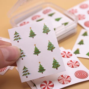 May include: A sheet of Christmas tree stickers with a white background and green trees. The sheet is being held by a hand. There are also sheets of candy cane stickers with red and white candy canes.