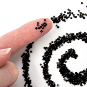May include: A close-up of a fingertip holding a small pile of black beads. A larger pile of black beads is arranged in a spiral pattern on a white surface.