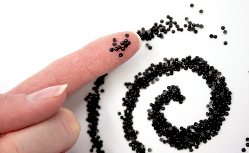 May include: A close-up of a fingertip holding a small pile of black beads. A larger pile of black beads is arranged in a spiral pattern on a white surface.