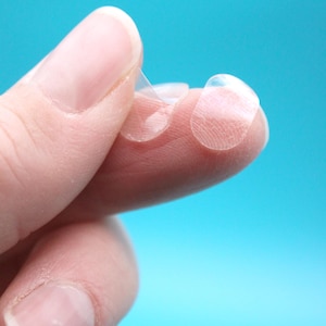 May include: Close-up of clear, adhesive nail protectors held between fingers against a bright blue background. The protectors are small, round, and transparent, designed to fit over the natural nail surface.