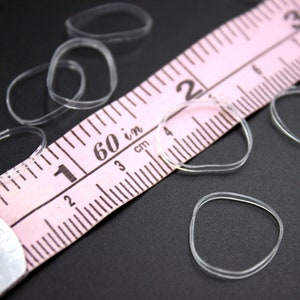 May include: A close-up of a pink measuring tape with clear rubber bands scattered around it. The tape measures in inches and centimeters. The tape is marked with numbers from 1 to 60.