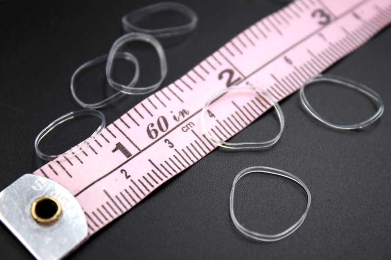 May include: A close-up of a pink measuring tape with clear rubber bands scattered around it. The tape measures in inches and centimeters. The tape is marked with numbers from 1 to 60.