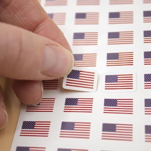 May include: A sheet of small stickers featuring the American flag. Each sticker has a white border, a blue rectangle with white stars, and red and white stripes. A hand is peeling one sticker from the sheet.