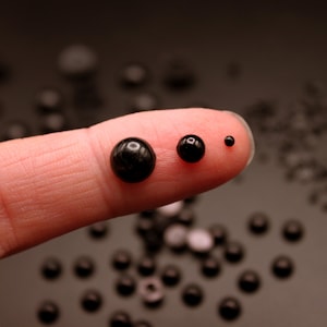 May include: A fingertip displays three black beads of varying sizes. The beads are round and smooth, with a glossy finish. The beads are arranged in order of size, with the largest bead on the left and the smallest bead on the right. The beads are surrounded by a scattering of smaller black beads.