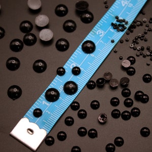 May include: A blue measuring tape with black markings lies on a dark surface with a scattering of black round beads and small black buttons.