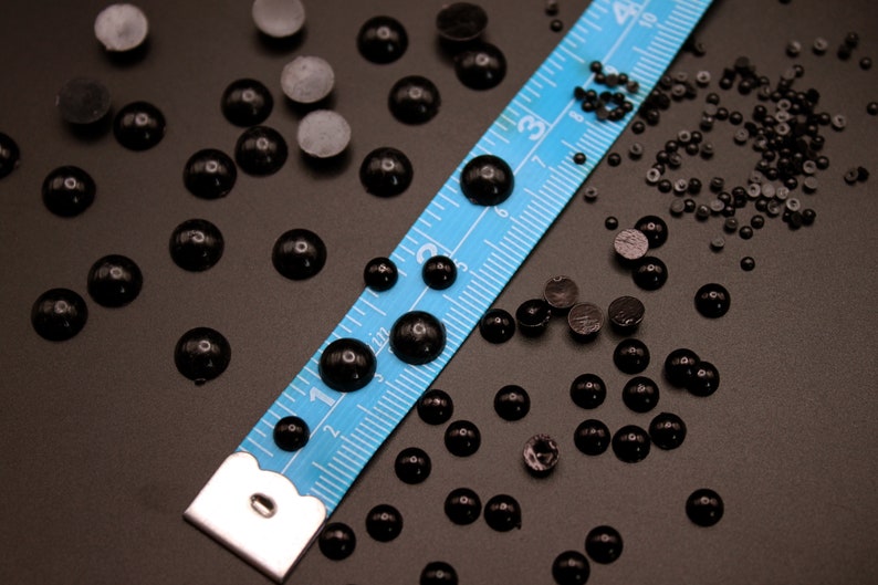 May include: A blue measuring tape with black markings lies on a dark surface with a scattering of black round beads and small black buttons.