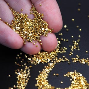May include: Close-up of a hand holding a pile of small, shiny, golden beads. The beads are scattered across a dark surface, reflecting light. The beads vary in size and shape, creating a textured appearance.