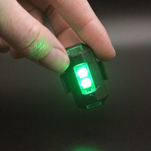 May include: A small, black, rectangular LED light with two bright green lights illuminated. The light is held between fingers against a dark background. The light is emitting a green glow.