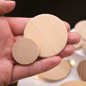 May include: A hand holds two unfinished, round wooden discs of different sizes, with a natural wood grain. Additional discs are visible in the background. These craft supplies are ideal for DIY projects, such as coasters or ornaments.