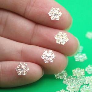 May include: Close-up of several iridescent snowflake-shaped embellishments. The small, decorative pieces are scattered on a green surface, with some resting on a fingertip. These are ideal for craft projects.