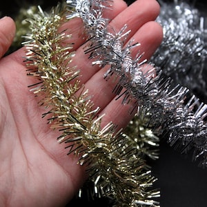 May include: Three strands of metallic tinsel garland in gold, silver, and a lighter silver color. The garland is made of thin, shiny strands of wire with small, pointed tips.