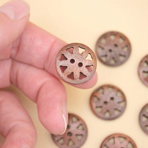 May include: A close-up of several brown wooden buttons. One button is held by a hand. The buttons are round with a decorative cut-out design and two small holes for sewing. The buttons are approximately 1 inch in diameter.