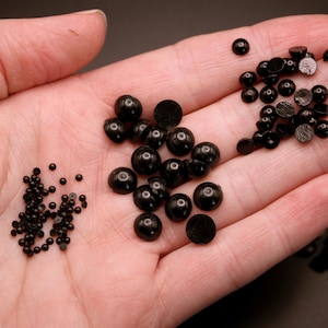 May include: A close-up of a hand holding three different sizes of black round plastic beads. The beads are arranged in three piles.