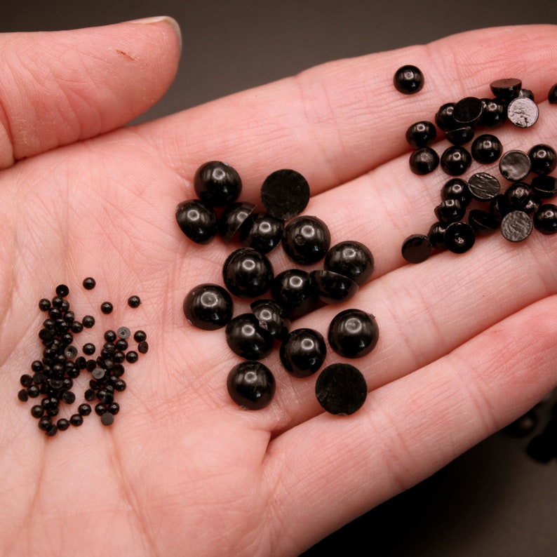 May include: A close-up of a hand holding three different sizes of black round plastic beads. The beads are arranged in three piles.