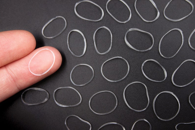 May include: A close-up of a finger holding a clear rubber band. The finger is surrounded by many other clear rubber bands on a black surface.