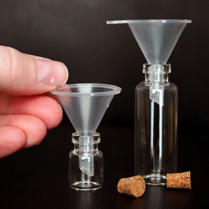 May include: Two clear plastic funnels with a glass bottle and cork stopper. The smaller funnel is being held by a hand. The larger funnel is sitting on top of a glass bottle with a cork stopper.