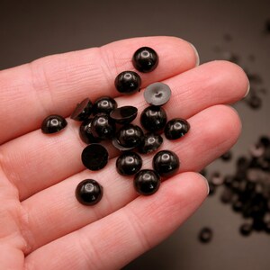 May include: A hand holding a pile of black plastic doll eyes. The eyes are round and have a shiny finish. There are more black plastic doll eyes scattered on a brown surface.
