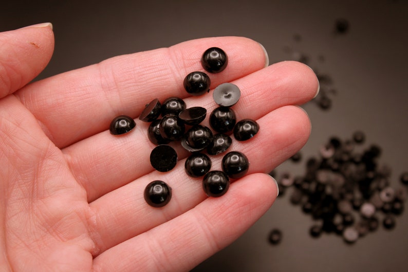 May include: A hand holding a pile of black plastic doll eyes. The eyes are round and have a shiny finish. There are more black plastic doll eyes scattered on a brown surface.
