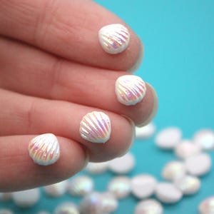May include: Small, white seashell-shaped beads with iridescent highlights are held against a blue background. The beads have a textured surface with radiating lines. Additional beads are scattered in the background.