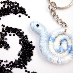 May include: A blue and white yarn keychain shaped like a snail with a black eye. The keychain is attached to a silver key ring. There are black beads scattered around the snail.