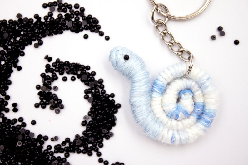 May include: A blue and white yarn keychain shaped like a snail with a black eye. The keychain is attached to a silver key ring. There are black beads scattered around the snail.