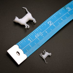 Puede incluir: Dos figuritas de gato de pl&aacute;stico blanco junto a una cinta m&eacute;trica azul con marcas blancas. La cinta mide hasta 7,6 cm.