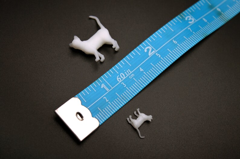 Puede incluir: Dos figuritas de gato de pl&aacute;stico blanco junto a una cinta m&eacute;trica azul con marcas blancas. La cinta mide hasta 7,6 cm.