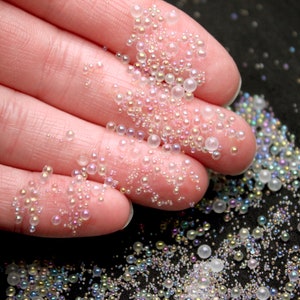 May include: Close-up of iridescent glass beads in various sizes, scattered on a black surface. The beads are held in a hand, showcasing their translucent, pearlescent quality. The beads reflect a rainbow of colors, creating a shimmering effect.