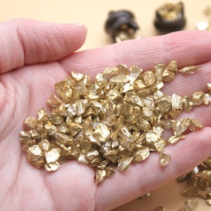 May include: A hand holding a pile of small, gold-colored, irregular shaped glass pieces. The pieces are scattered on a light brown surface.