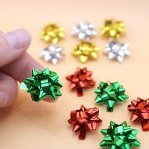 May include: A collection of small, shiny, metallic gift bows in various colors including green, red, gold, and silver. The bows are star-shaped with multiple loops and a reflective finish, arranged in a group with one green bow held up.