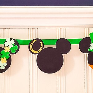 May include: A green and black banner featuring Mickey Mouse heads decorated with St. Patrick's Day themed elements such as shamrocks, rainbows, and gold coins.
