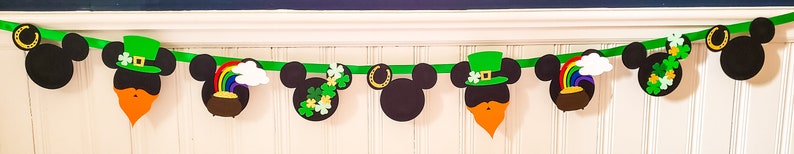 May include: A green and black banner featuring Mickey Mouse heads decorated with St. Patrick's Day themed elements such as shamrocks, rainbows, and gold coins.