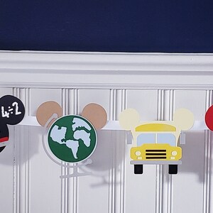 May include: A colorful banner with Mickey Mouse ears features school-themed images, including a school bus, an apple, pencils, a globe, and a chalkboard with the letters "ABC" and the numbers "1+2".