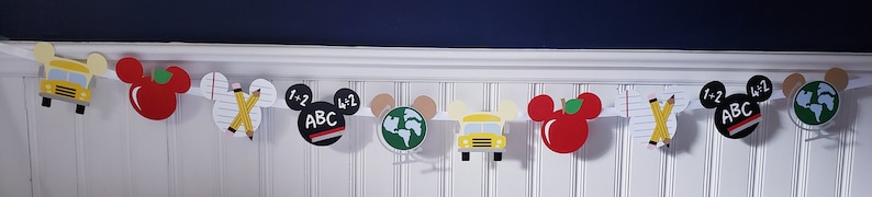 May include: A colorful banner with Mickey Mouse ears features school-themed images, including a school bus, an apple, pencils, a globe, and a chalkboard with the letters "ABC" and the numbers "1+2".