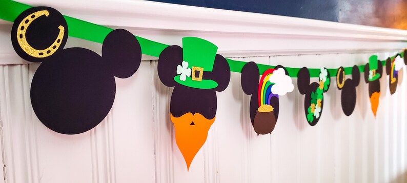 May include: A green, white and gold St. Patrick's Day banner featuring Mickey Mouse heads with various Irish-themed decorations, including a pot of gold, a rainbow, a shamrock and a leprechaun hat.