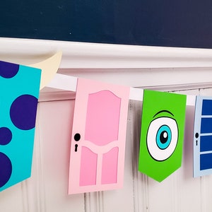 May include: A colourful paper banner featuring characters from the film Monsters Inc. The banner includes a blue and purple monster with horns, a pink door, a green monster with one eye, a blue door, a purple monster with hair, a white door with flowers, and a blue monster with one eye.