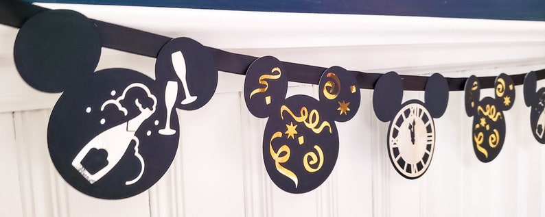 May include: Black and gold New Year's Eve party banner with Mickey Mouse ears and different designs, including a champagne bottle, a clock, and confetti.