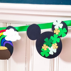 May include: A green, black and gold Mickey Mouse themed banner featuring shamrocks, a pot of gold, a rainbow, a leprechaun hat and a horseshoe. The banner is made of paper and is ideal for a St. Patrick's Day party.