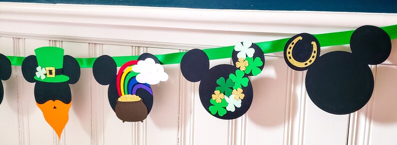 May include: A green, black and gold Mickey Mouse themed banner featuring shamrocks, a pot of gold, a rainbow, a leprechaun hat and a horseshoe. The banner is made of paper and is ideal for a St. Patrick's Day party.
