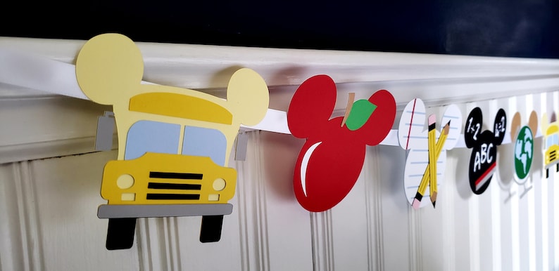May include: A back-to-school themed banner with Mickey Mouse ears. The banner features a yellow school bus, a red apple, a sheet of paper with pencils, a Mickey Mouse head with the letters "ABC" and the numbers "1+2", a globe, and a yellow school bus.