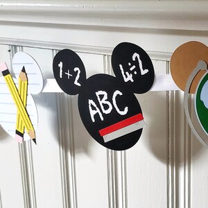 May include: A white banner with Mickey Mouse ears features various school-themed images, including a school bus, apple, pencils, a chalkboard with the text "1+2" and "4:2", a chalkboard with the text "ABC", and a globe.